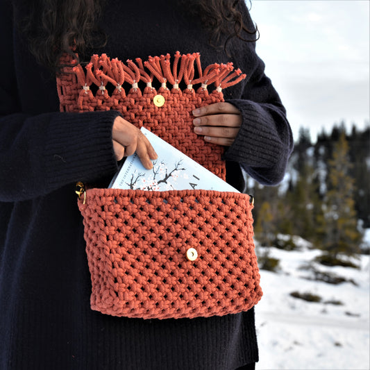 A woman taking a book out of a brick coloured woven shoulder bag with fringes and a magnetic clasp