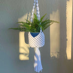 Load image into Gallery viewer, A white basket-style macrame plant hanger with a fern plant  in a blue planter.
