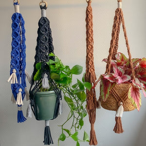 A blue macrame plant hanger with white fringe detailing, a black macrame plant hanger with grey fringe detailing and two brown macrame plant hangers with brown and white fringe detailing hanging from a rod