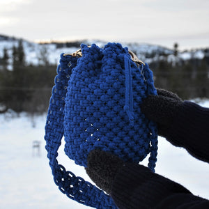 A royal blue woven macrame bucket bag with drawstring closure and a shoulder strap