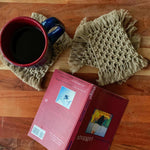 Load image into Gallery viewer, A set of 4 beige macrame coasters with a cup of tea on a coaster and a book near it on the table.
