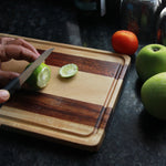 Load image into Gallery viewer, A lemon being cut on a walnut wood and birch wood striped chopping board
