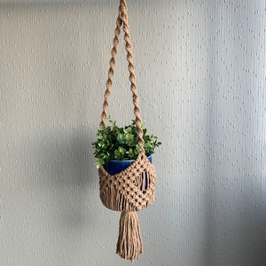 A brown macrame basket plant hanger with a green plant in a blue planter hanging from the ceiling