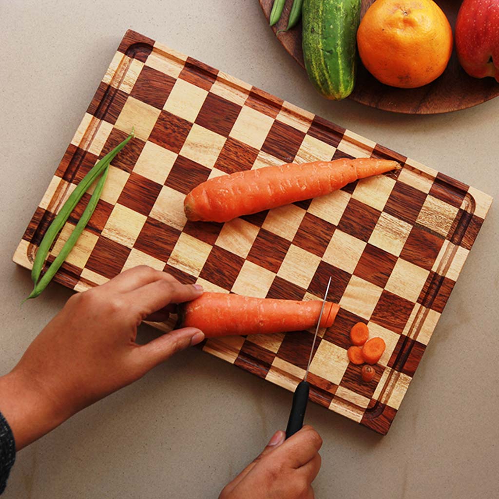 A chessboard style wooden cutting board made with brown walnut and birch wood with carrots being cut on it.
